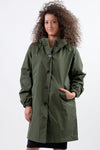 A person with curly hair is wearing an olive-green, lightweight, water-resistant jacket with an adjustable hood and their left hand placed in the pocket. The OLIVE GREEN WINDBREAKER from The Windbreaker Jacket features a button-up design and the person is standing against a plain, light-colored background.