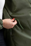 A person with a red manicure adjusts the drawstring on an olive green, lightweight water-resistant OLIVE GREEN WINDBREAKER from The Windbreaker Jacket. Only the hand, part of the arm, and the lower torso are visible. They are wearing a white shirt and dark pants.