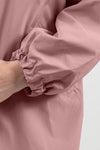 Close-up of an arm wearing a The Windbreaker Jacket PASTEL PINK WINDBREAKER with elasticated cuffs. The sleeve is slightly gathered at the wrist, showcasing the smooth texture and stitching details of this stylish water-resistant jacket. The background is a plain off-white color.