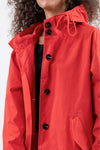 A person with curly hair wearing a stylish CRIMSON RED WINDBREAKER by looselyboho with black buttons is shown from the shoulders up. The water-resistant jacket features a drawstring hood and a pocket on the side. The background is a plain, light-colored backdrop.